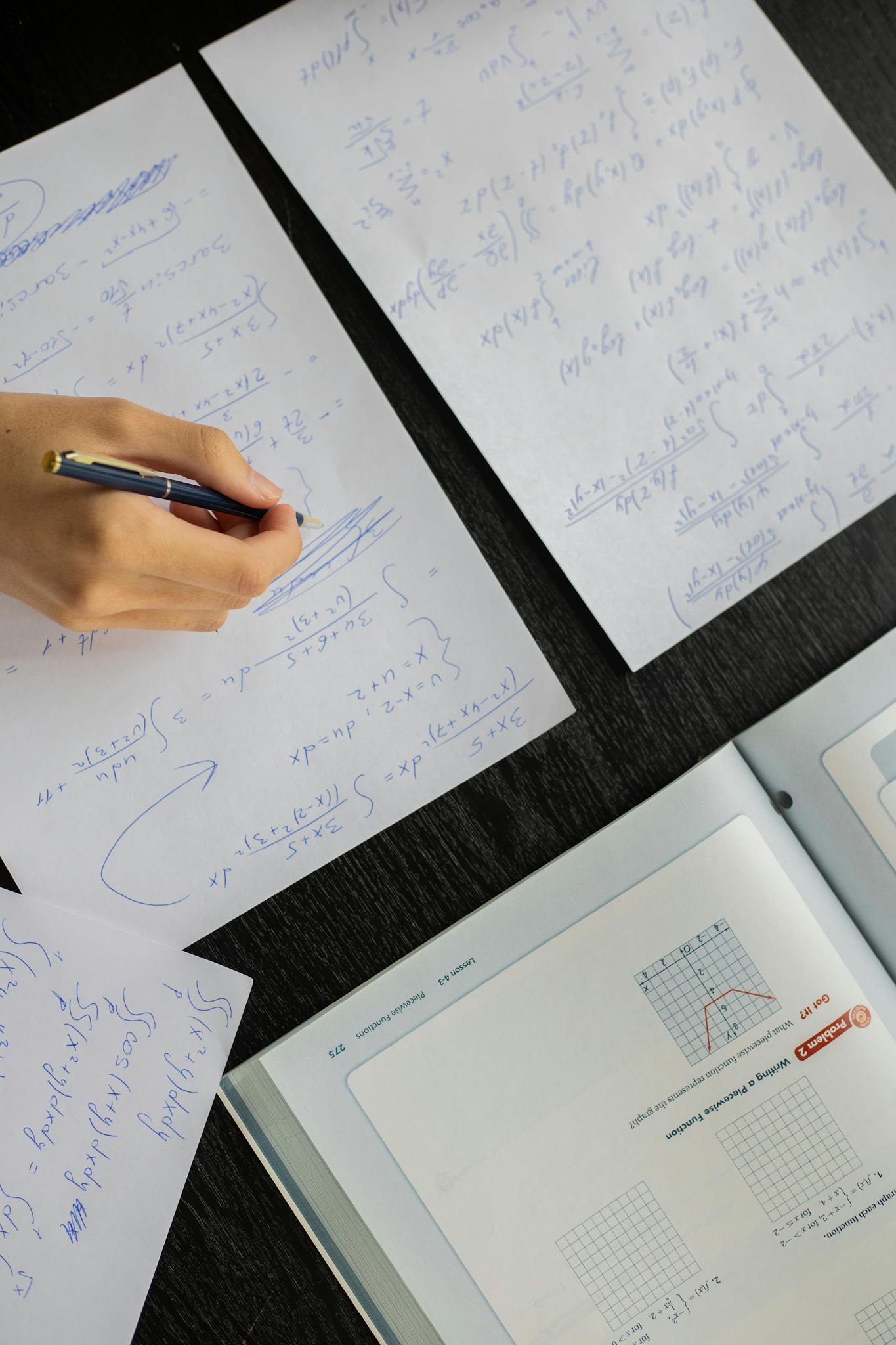 Close-up of a student solving mathematical equations with papers and textbook on a table.